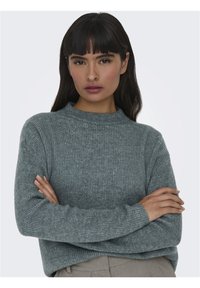 Pull gris côtelé avec col montant et manches longues, présentant un motif de tricot texturé. Le mannequin pose les bras croisés.