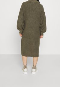 Stickad klänning i olivgrön med hög krage och oversized ärmar, knälång. Bärs med vita sneakers och mönstrade strumpor.