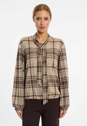 Frau trägt eine beige und braun karierte langärmelige Bluse mit Schlupfkragen und dunkelbraune Hose, steht vor einem weißen Hintergrund.