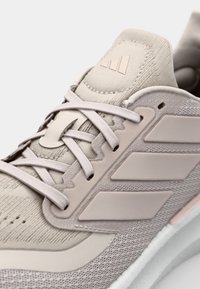 Chaussure de sport beige clair avec une tige en tissu texturé, logo à trois bandes, fermeture à lacets et semelle intermédiaire blanche rembourrée.