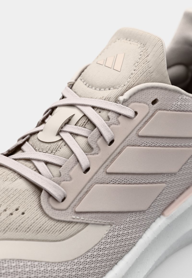 Chaussure de sport beige clair avec une tige en tissu texturé, logo à trois bandes, fermeture à lacets et semelle intermédiaire blanche rembourrée.