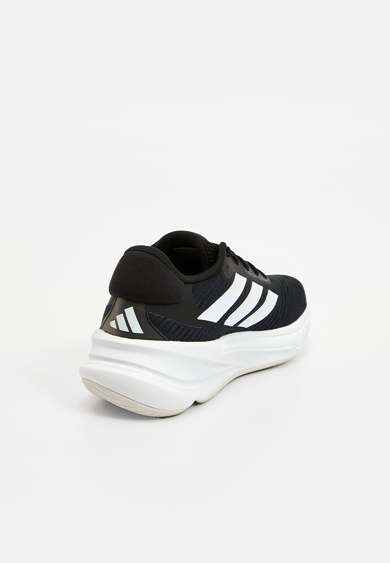 Scarpa sportiva nera con un rivestimento testurizzato, strisce bianche di accento e suola ammortizzata. Presenta una forma arrotondata e un colletto imbottito.