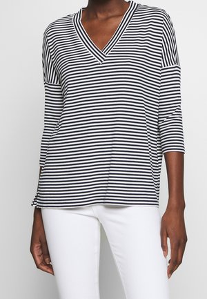 Personne portant un haut à col en V rayé noir et blanc à manches trois-quarts et un pantalon blanc, debout devant un fond uni.