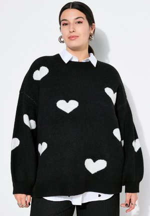 Maglione nero in morbido maglione, con motivi a cuore bianchi. Vestibilità ampia con orlo a coste. Indossato sopra una camicia bianca con colletto.