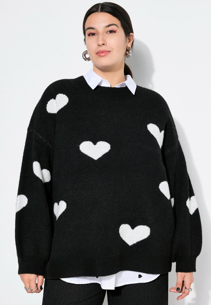 Schwarzer Pullover aus weichem Strick, mit weißen Herzmustern. Lockere Passform mit ribbed Saum. Über einem weißen Hemd mit Kragen getragen.