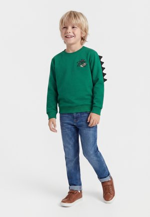 Jeune garçon souriant portant un sweat vert avec un patch dragon et des pointes noires dans le dos, jean bleu et baskets marron.