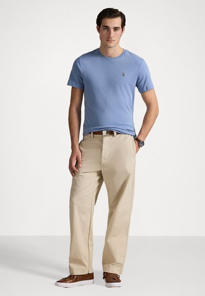 Heleheles sinine ümar kaelusega t-särk, beežid chinos-püksid, pruunid nahast tossud ja pruun vöö. T-särgil on väike logo. Lihtne disain.