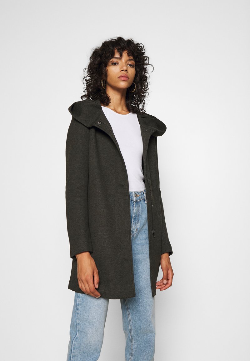 Manteau vert foncé avec une capuche, doté d'une fermeture éclair, à manches longues et à coupe décontractée, associé à un jean taille haute bleu clair.