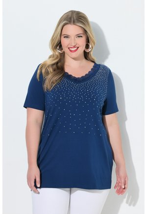 Glimlachende vrouw met lang blond haar, grote hoepeloorbellen, een marineblauwe versierde V-hals top en witte broek tegen een effen achtergrond.