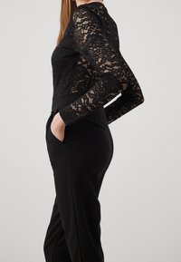 Svart spetsbodysuit med långa ärmar, i kombination med svarta skräddarsydda byxor med fickor, som framhäver intrikata blommönster i spetsen.