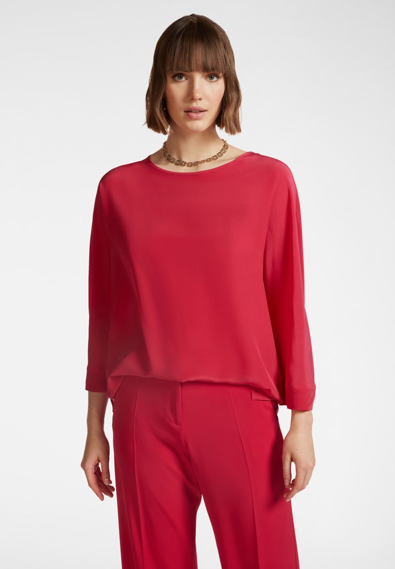 Blusa rossa realizzata in un tessuto leggero e liscio. Presenta un collo rotondo e maniche lunghe, abbinata a pantaloni rossi coordinati.