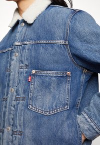 Jeansjacka med ljusblå tvätt, fårskinnskrage, koppardetaljer, synlig sömnad och en enfaldig bröstficka med en logotagg.