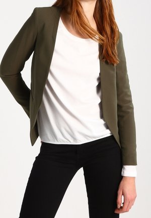 Osoba nosząca otwartą oliwkowo-zieloną marynarkę na białej koszuli z długim rękawem i czarnych spodniach, stojąca z jedną ręką na biodrze.