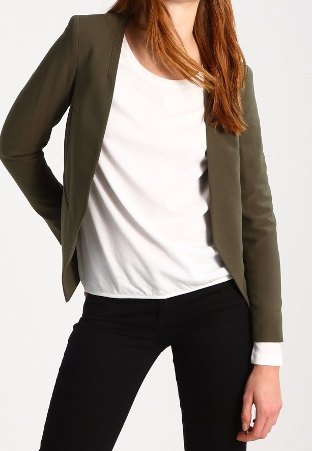 Personne portant un blazer vert olive ouvert sur une chemise blanche à manches longues et un pantalon noir, debout avec une main sur la hanche.