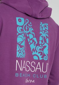 Violetti huppari, jossa on suuri sininen koristeellinen "N", joka on täytetty trooppisilla kuvioilla, ja "NASSAU BEACH CLUB" -teksti valkoisena alapuolella.