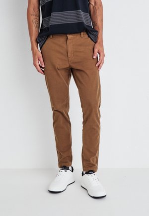 Man met bruine slim-fit broek, witte sneakers en een zwart shirt met patroon, staand tegen een effen lichte achtergrond.