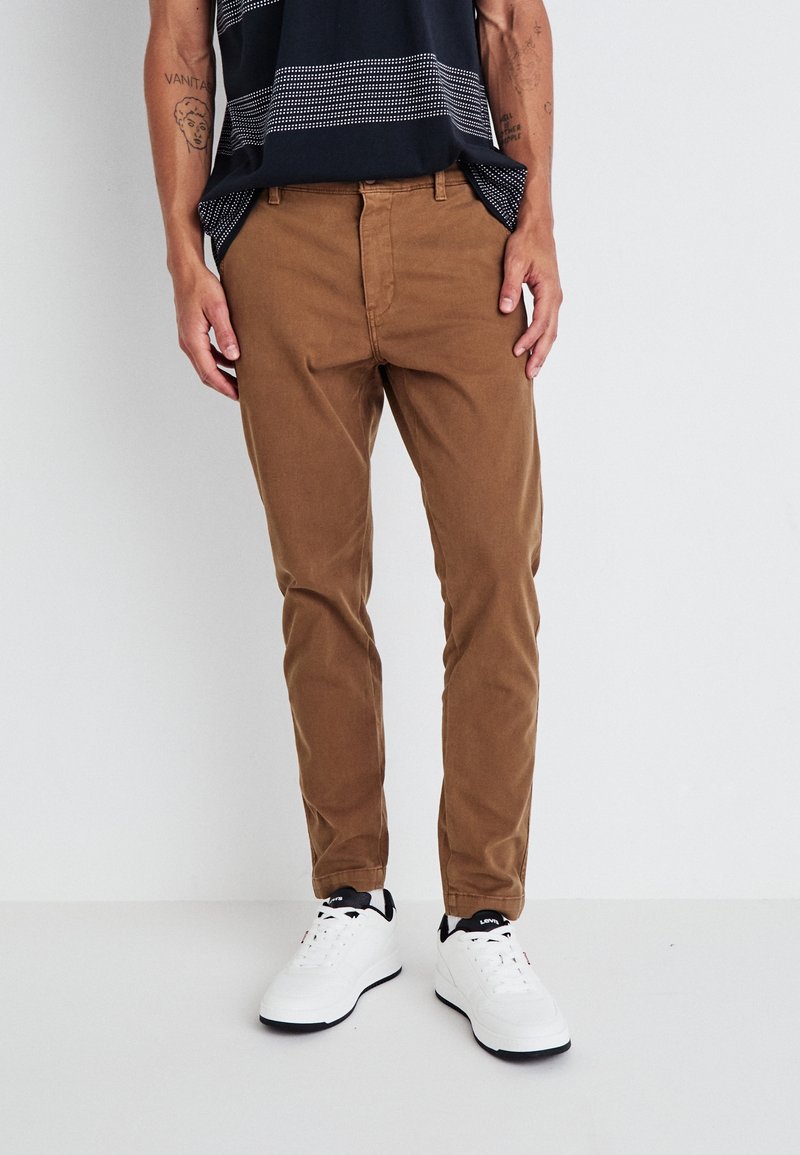 Homme portant un pantalon marron slim, des baskets blanches et une chemise noire à motifs, debout devant un fond clair uni.