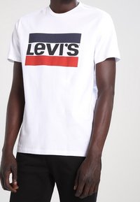 Vit kortärmad t-shirt med en röd och marinblå Levi's-logotyp tvärs över bröstet. Tillverkad av bomull med en avslappnad passform.
