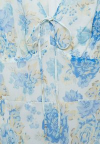 Blumenkleid aus leicht blauem, transparentem Stoff mit weißen und beigen Blumen, einer Bindeschließe und weicher, fließender Textur.