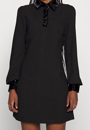 Kvinde iført en sort langærmet kjole med skinnende sort krave og manchetter, stående mod en ensfarvet lys baggrund.