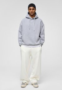 Grijze sweatshirt met een capuchon, grote voork pocket, losse pasvorm, gecombineerd met witte pantalon met wijde pijpen en grijze sneakers met gaasaccenten.