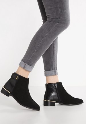 Zwarte enkellaarzen met gouden accenten gedragen bij opgerolde grijze skinny jeans, getoond met één voet licht omhoog en de andere plat op de grond.