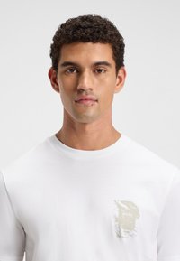 Tricou alb din bumbac cu maneci scurte, prezentând un design de logo estompat în bej în zona din stânga a pieptului. Textură netedă și guler rotund.