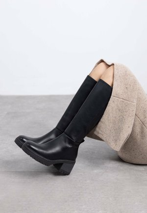 Botas de cuero negro hasta la rodilla con punta redonda y suela texturizada, que cuentan con un tacón cuadrado moderado y un diseño vertical sin costuras.