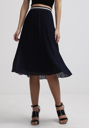 Faltenrock in Marineblau in Midi-Länge mit strukturierter Oberfläche und kontrastierendem Bund. Gestylt mit schwarzen Riemchensandalen.