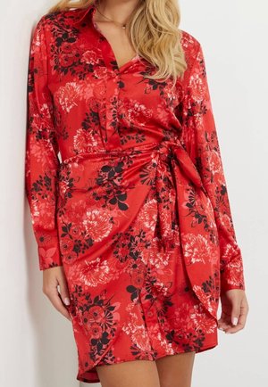 Femme portant une robe portefeuille rouge à motifs floraux noirs et roses, manches longues et boutonnage devant.