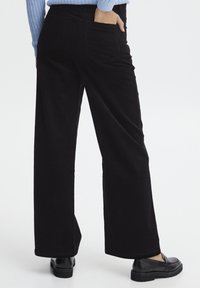 Zwarte corduroy wijde pijpen broek met een gestructureerd oppervlak, voorzien van achterzakken en een middelhoge taille, gecombineerd met glanzende zwarte instappers.