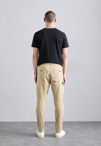Zwarte t-shirt met korte mouwen, beige broek, beide in een relaxte pasvorm. De broek heeft zijzakken en een gladde stoftextuur. Witte sneakers.