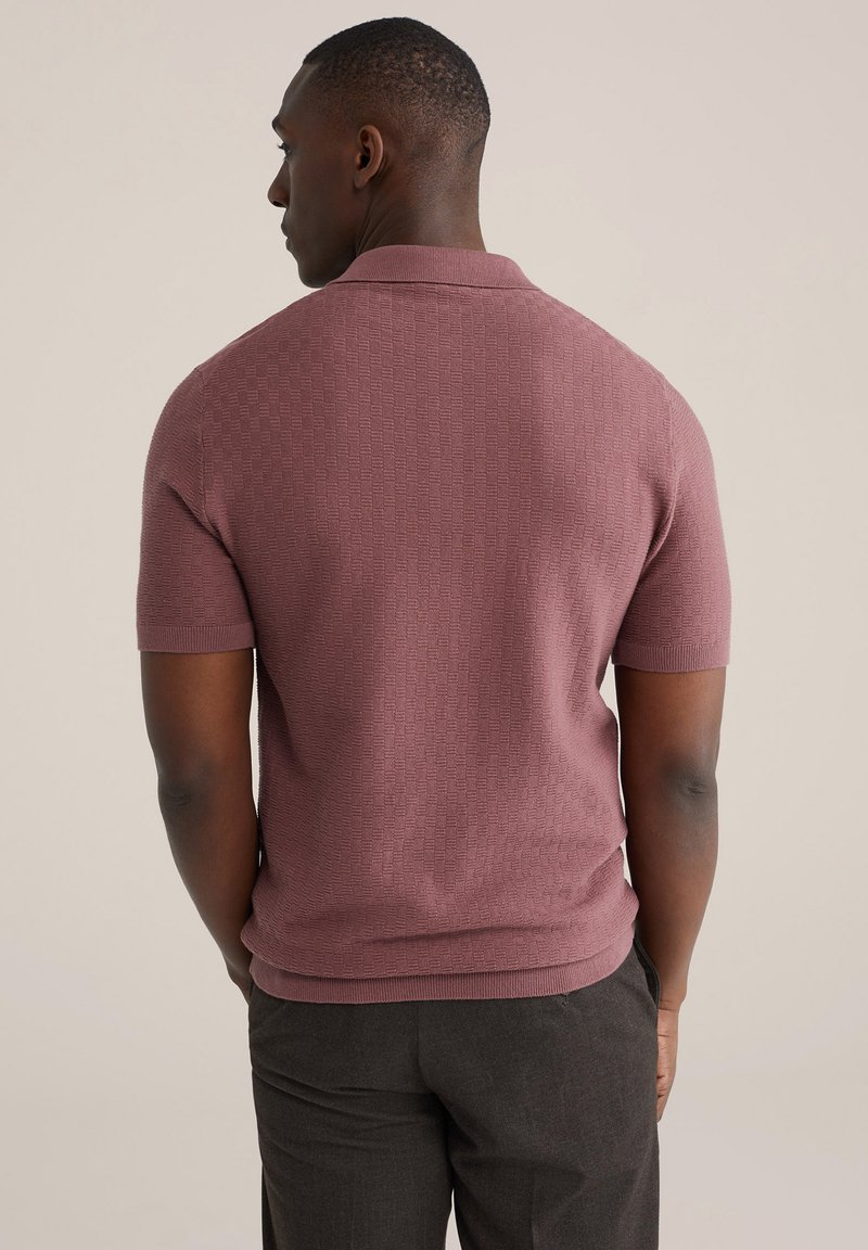 Polo à manches courtes couleur rose poudré, présentant un motif en maille texturée et un col classique. Porté avec un pantalon sombre ajusté.