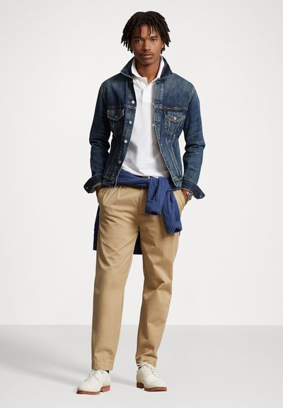 Junger Mann steht mit den Händen in den Taschen, trägt eine Jeansjacke, weißes Polo, beigefarbene Hose, weiße Schuhe und einen blauen Pullover, der um die Taille gebunden ist.