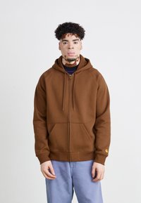 Sudadera con cremallera de color marrón, con un bolsillo tipo canguro, puños acanalados y un pequeño logo amarillo en la manga izquierda. Llevada sobre una camiseta negra.