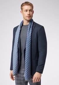 Marineblazer med struktureret mønster, grå sweater og et lyseblå tørklæde med geometrisk design og frynser. Bukserne er ternede.