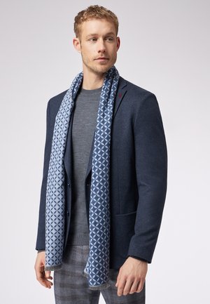 Marineblazer med struktureret mønster, grå sweater og et lyseblå tørklæde med geometrisk design og frynser. Bukserne er ternede.