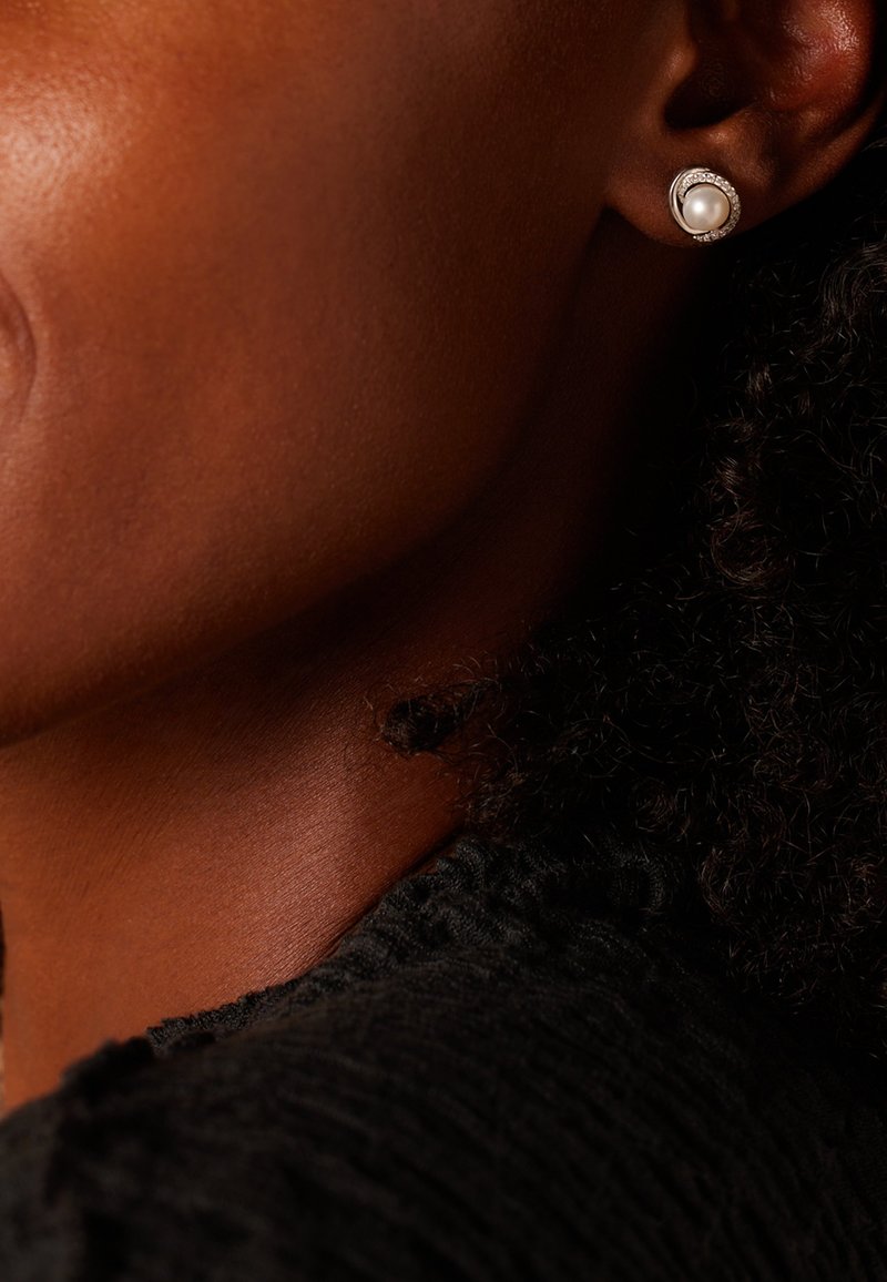 Boucle d'oreille en argent avec une perle ronde entourée de petites pierres claires. Vêtements noirs texturés partiellement visibles en arrière-plan.