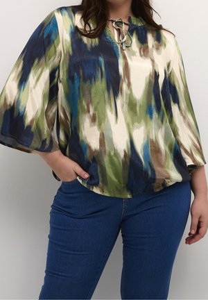 Kvinde iført en løs, langærmet bluse med et abstrakt penselstrøgsmønster i blå, grøn, creme og brun, kombineret med blå jeans.