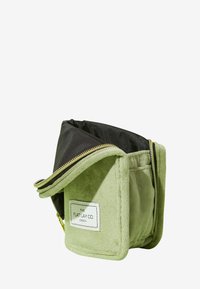 Grønn stoffoppbevaringspose med myk tekstur, svart innside og gullglidelåser. Har et merkelapp med "THE FLAT LAY CO. LONDON" på siden.