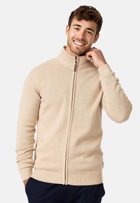 Beige ritsvest met een hoge geribde kraag en een textuur breisel. Het heeft geribde manchetten en een zoom, en is voorzien van een bruine ritstrekkertje.