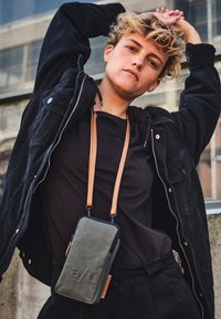 Black fabric jacket over a black shirt, paired with high-waisted trousers. A dark green phone bag with a tan strap hangs from the neck.