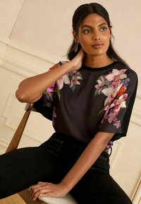 Donna con lunghi capelli scuri seduta, indossa un top nero con motivi floreali rosa e arancione, con una mano appoggiata al collo e l'altra in grembo.