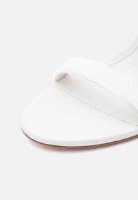Sandale blanche avec une large bride texturée unique sur la zone des orteils et une semelle plate à bord légèrement arrondi.
