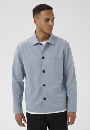 Világoskék, gombos dzsekit viselő férfi fehér ing fölött, sima háttér előtt áll, oldalra néz.