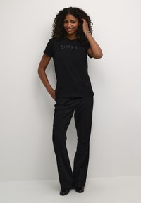 T-shirt en coton noir avec le texte "CHERISH" en matériau brillant, associé à un pantalon noir ajusté. Le mannequin pose les mains sur les hanches, souriant.