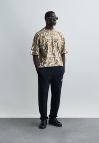 Άνδρας που φοράει καφέ πουκάμισο Moschino με σχέδια, μαύρο παντελόνι jogger, μαύρα αθλητικά παπούτσια και γυαλιά ηλίου, στέκεται με τα χέρια στις τσέπες.