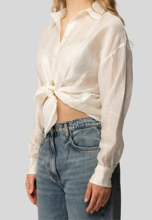 Donna che indossa una camicia bianca trasparente a maniche lunghe annodata in vita e jeans blu a vita alta su sfondo grigio.
