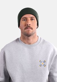 Bonnet en tricot vert, texture côtelée, porté avec un sweat-shirt gris clair orné de broderies florales décoratives en bleu et jaune.