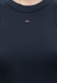 Κοντινό πλάνο ενός ατόμου που φοράει μπλε σκουρόχρωμη μπλούζα χωρίς μανίκια με ριμπ ύφανση και μικρό ορθογώνιο κόκκινο, λευκό και μπλε λογότυπο στο κέντρο του στήθους.